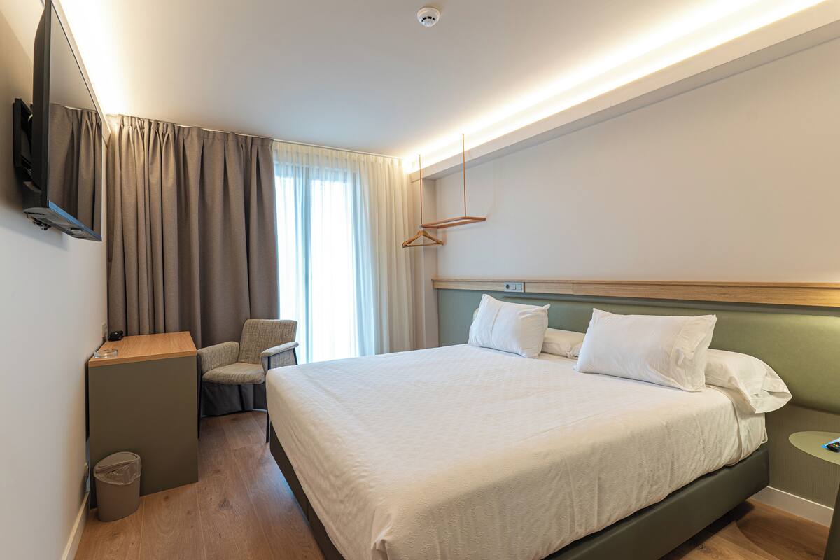 A hotel room with a bed, a chair, a small table, a TV, and a bedside table.