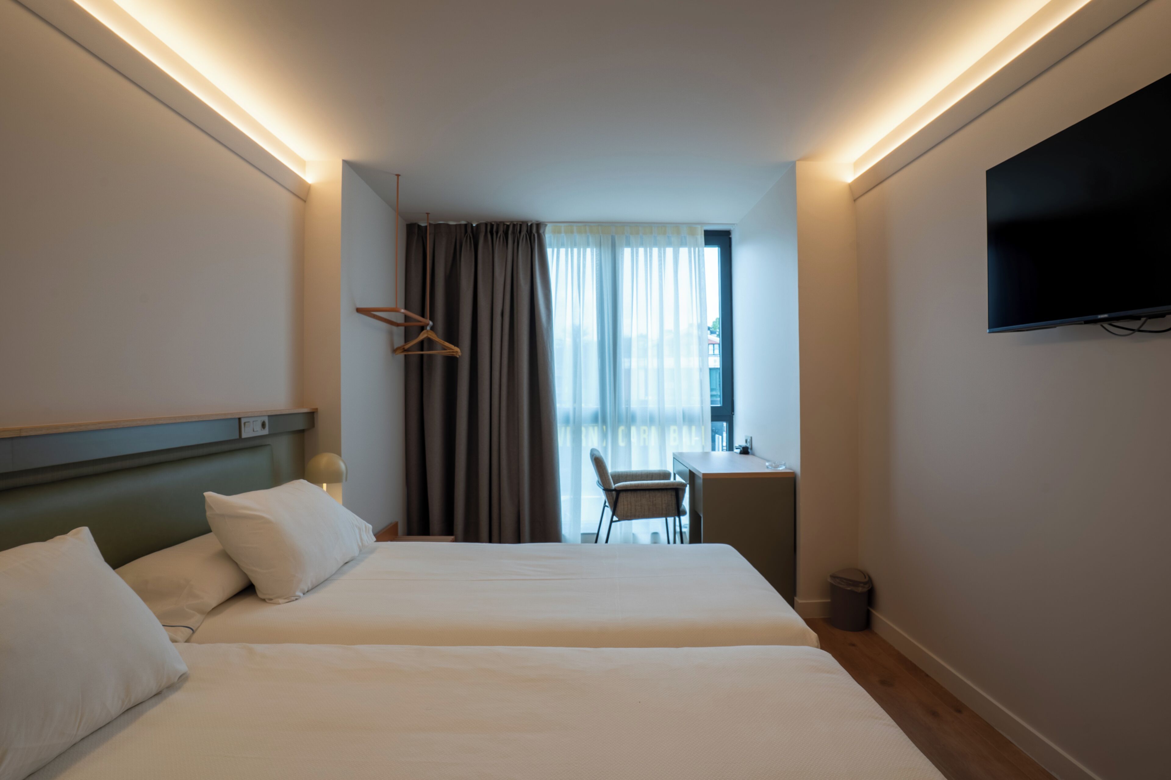 A hotel room with two beds, a desk, a chair, a TV, and a window with curtains.