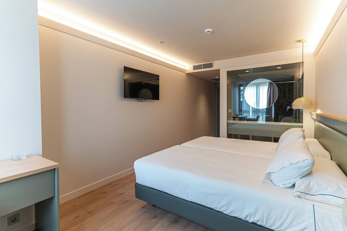 A modern hotel room with a bed, a TV, a mirror, and a bedside table.