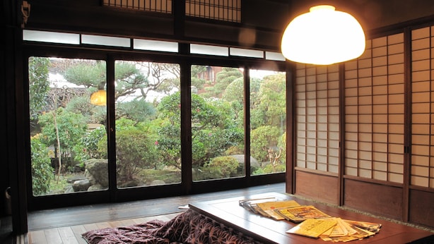 Ett traditionellt japanskt rum med tatamimattor på golvet, ett lågt bord med kuddar och utsikt över en trädgård genom stora fönster.