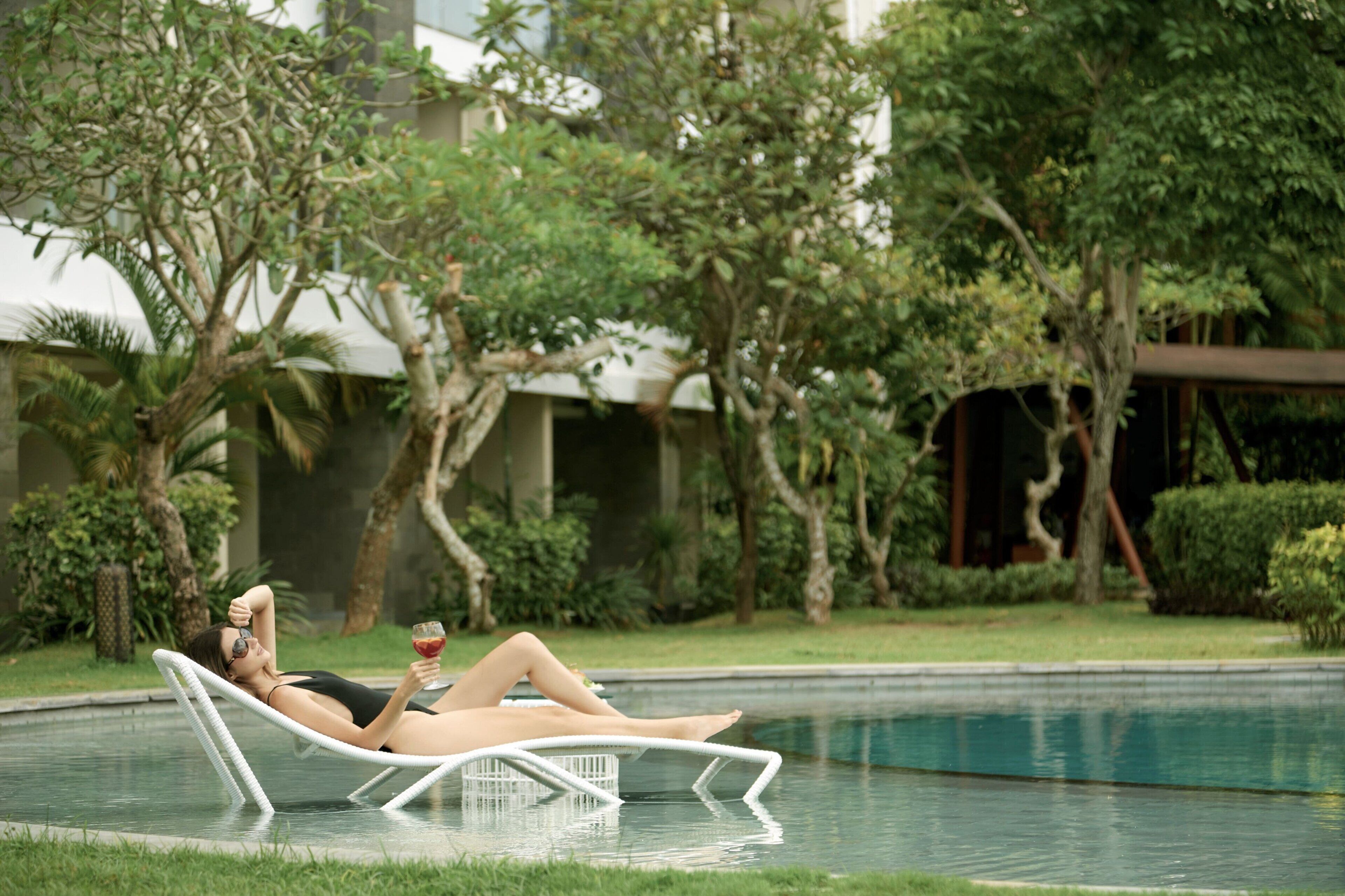 Una persona descansando en una silla reclinable blanca junto a una piscina, rodeada de follaje y un edificio al fondo.