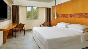 A hotel room with a large bed, a desk, two chairs, a TV, and a window with curtains.