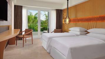 A hotel room with a desk, two beds, a chair, and a large window with a view of greenery.