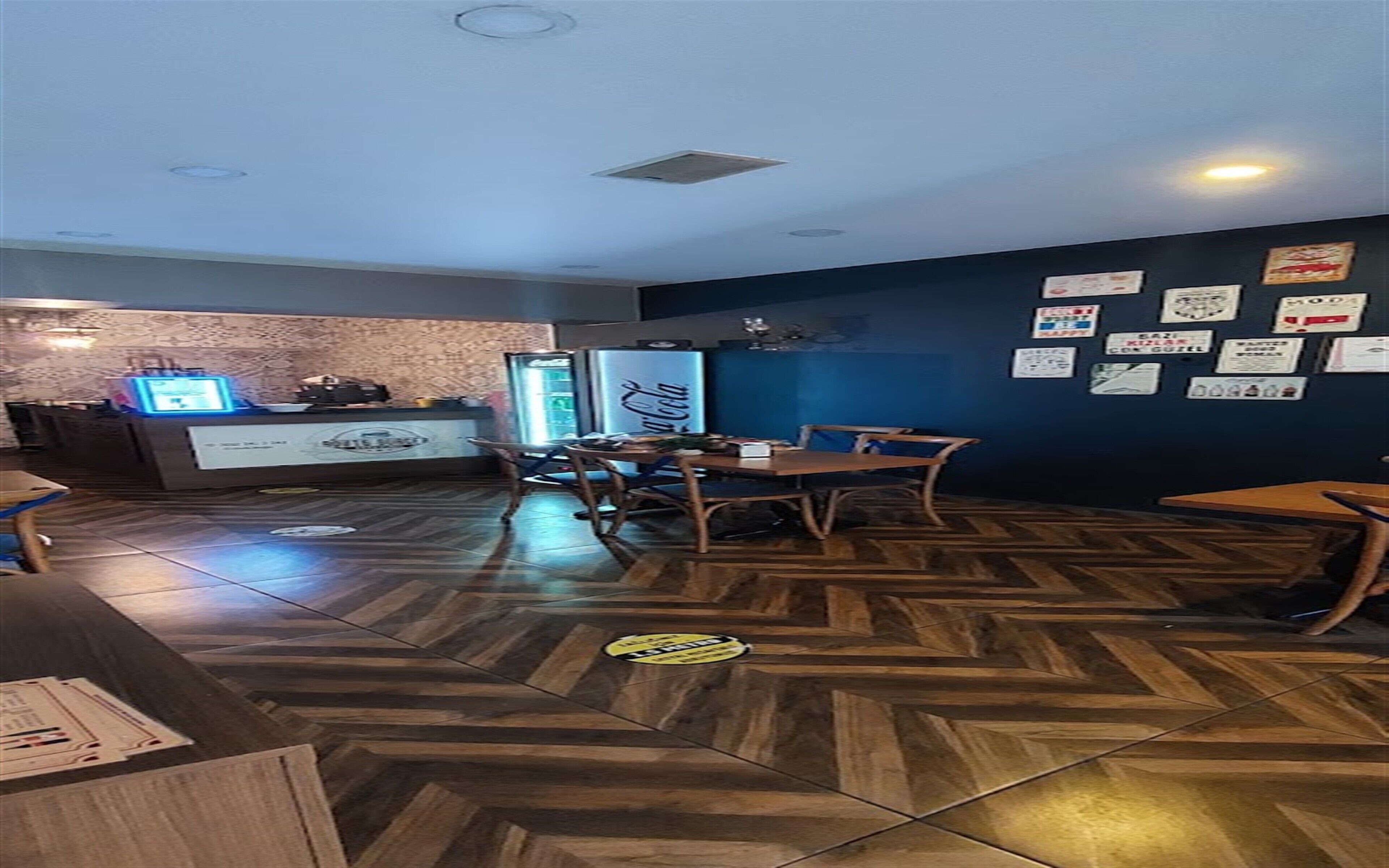 A restaurant with a herringbone-patterned floor, a counter with a computer and a refrigerator, and a wall decorated with various signs.