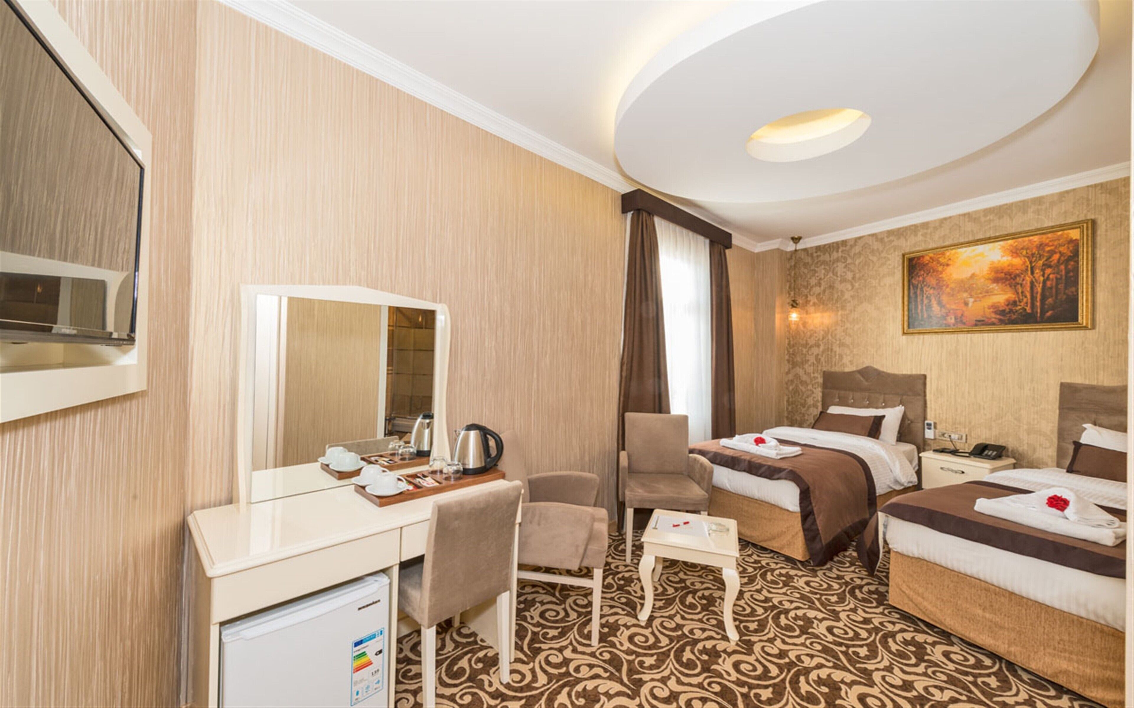 A hotel room with a desk, chair, two beds, a mirror, a painting, and a patterned carpet.