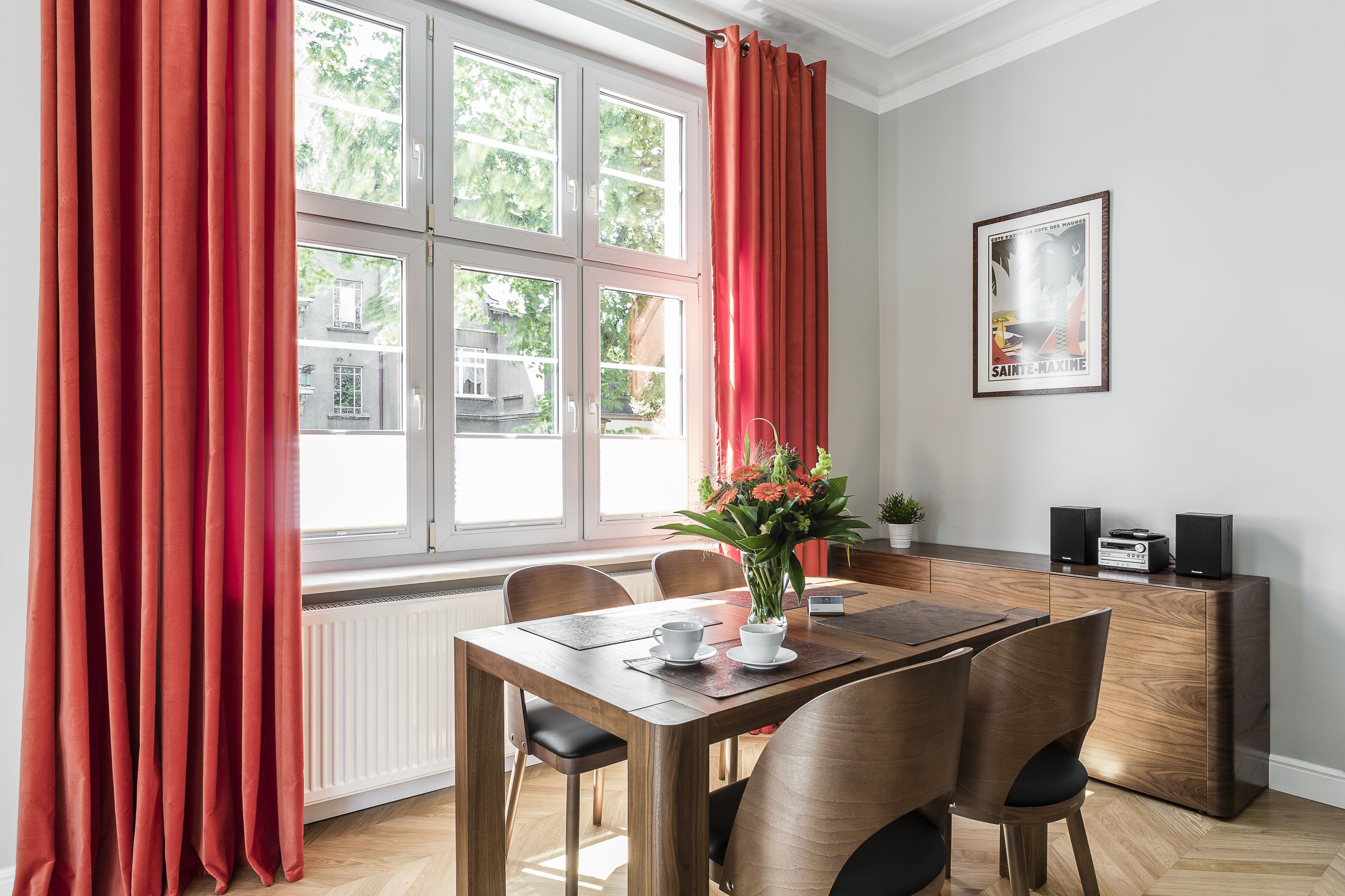 A dining area with a wooden table, chairs, a vase with flowers, and a radiator below the window.