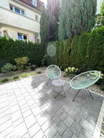 Un patio con dos sillas de malla metálica verde y una pequeña mesa redonda, rodeado de un seto bien cuidado y un edificio al fondo.