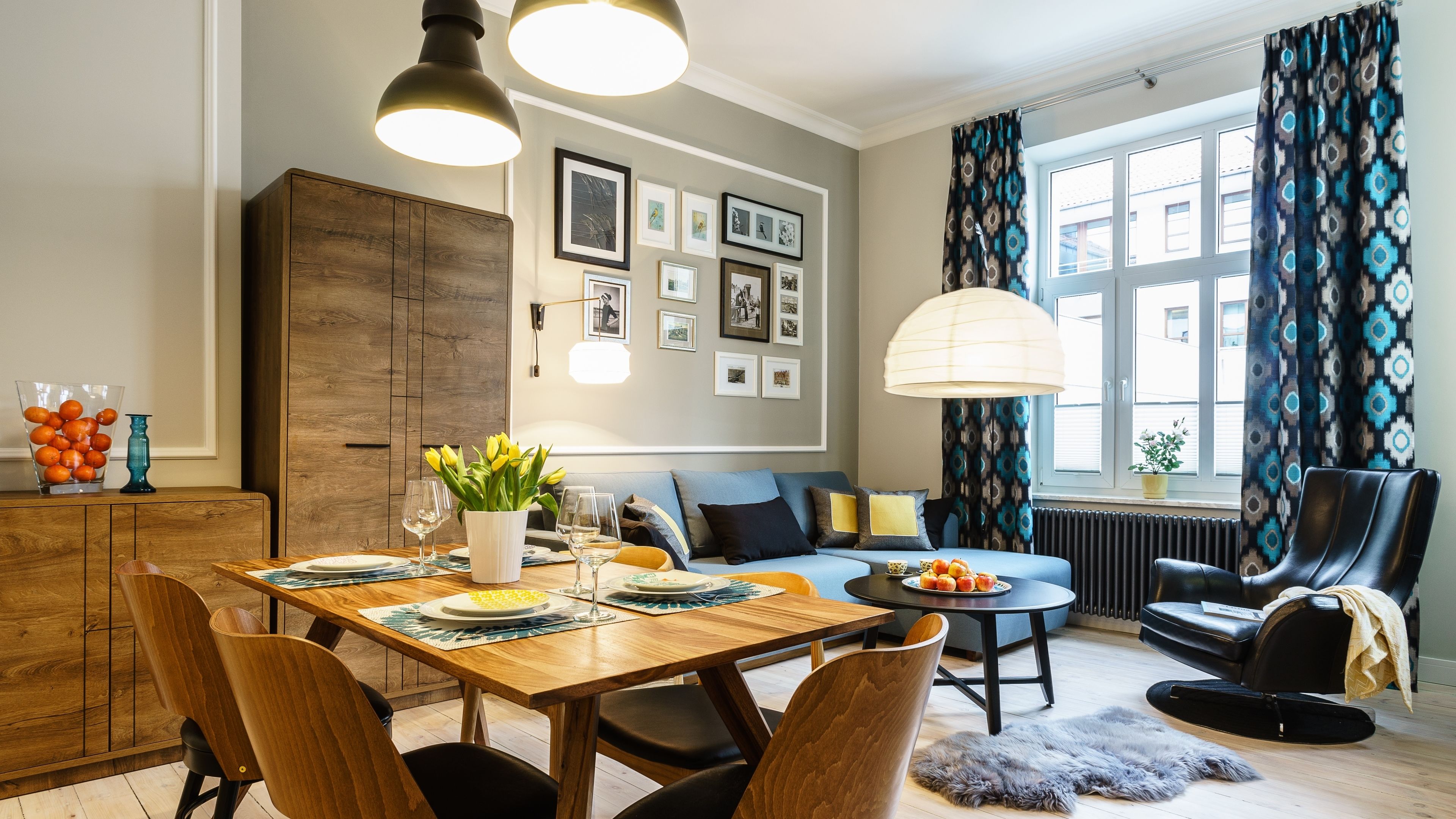 A dining area with a wooden table set for four, surrounded by chairs. There is a sofa and a chair in the background, and a window with patterned curtains.