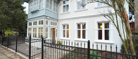 A two-story house with a black iron fence, a stone pathway, and a garden with trees and shrubs.