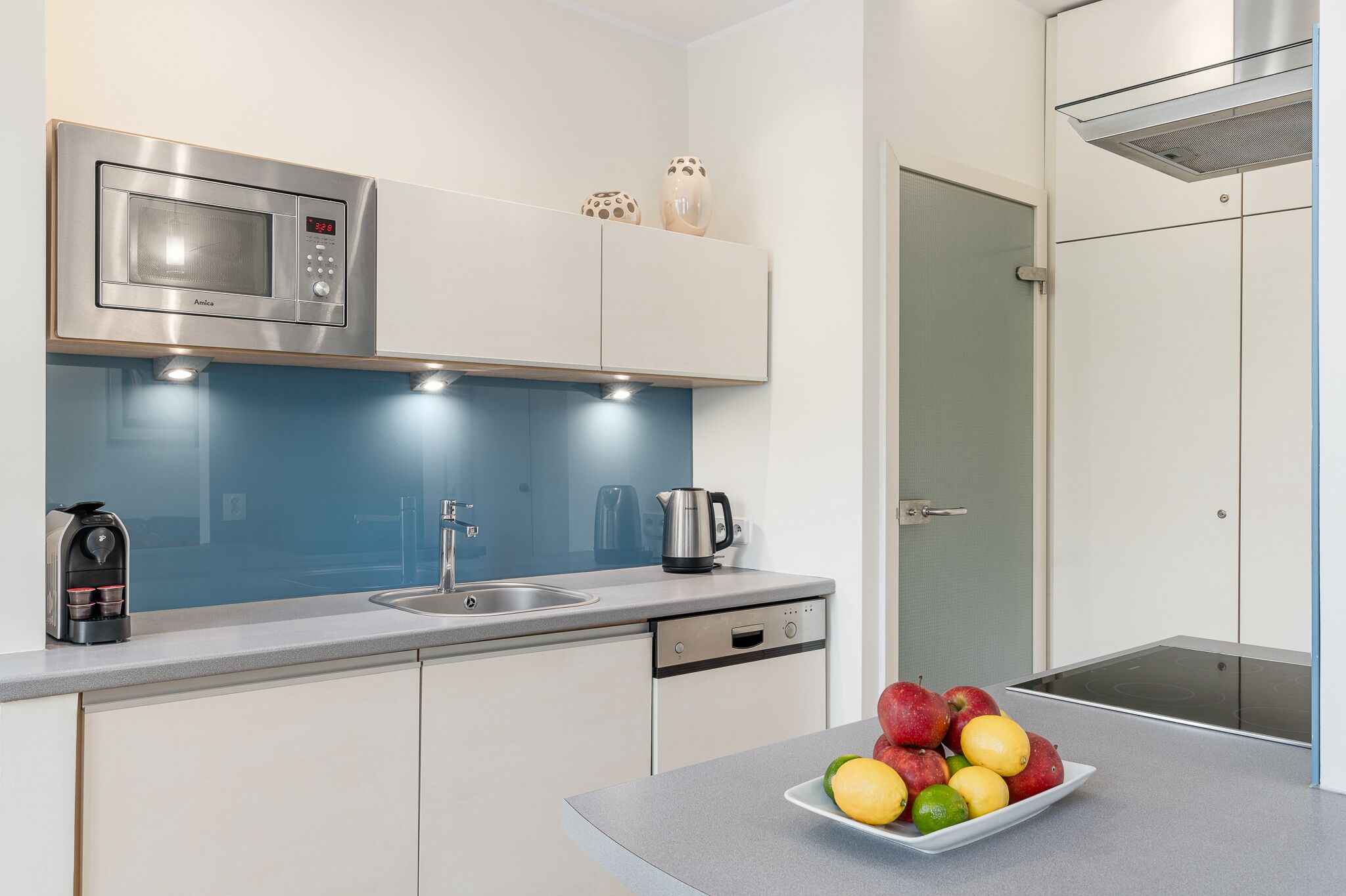 A modern kitchen with a built-in microwave, a coffee maker, and a kettle on the counter. There is a sink and a dishwasher below the counter. A plate of fruit is on the counter.