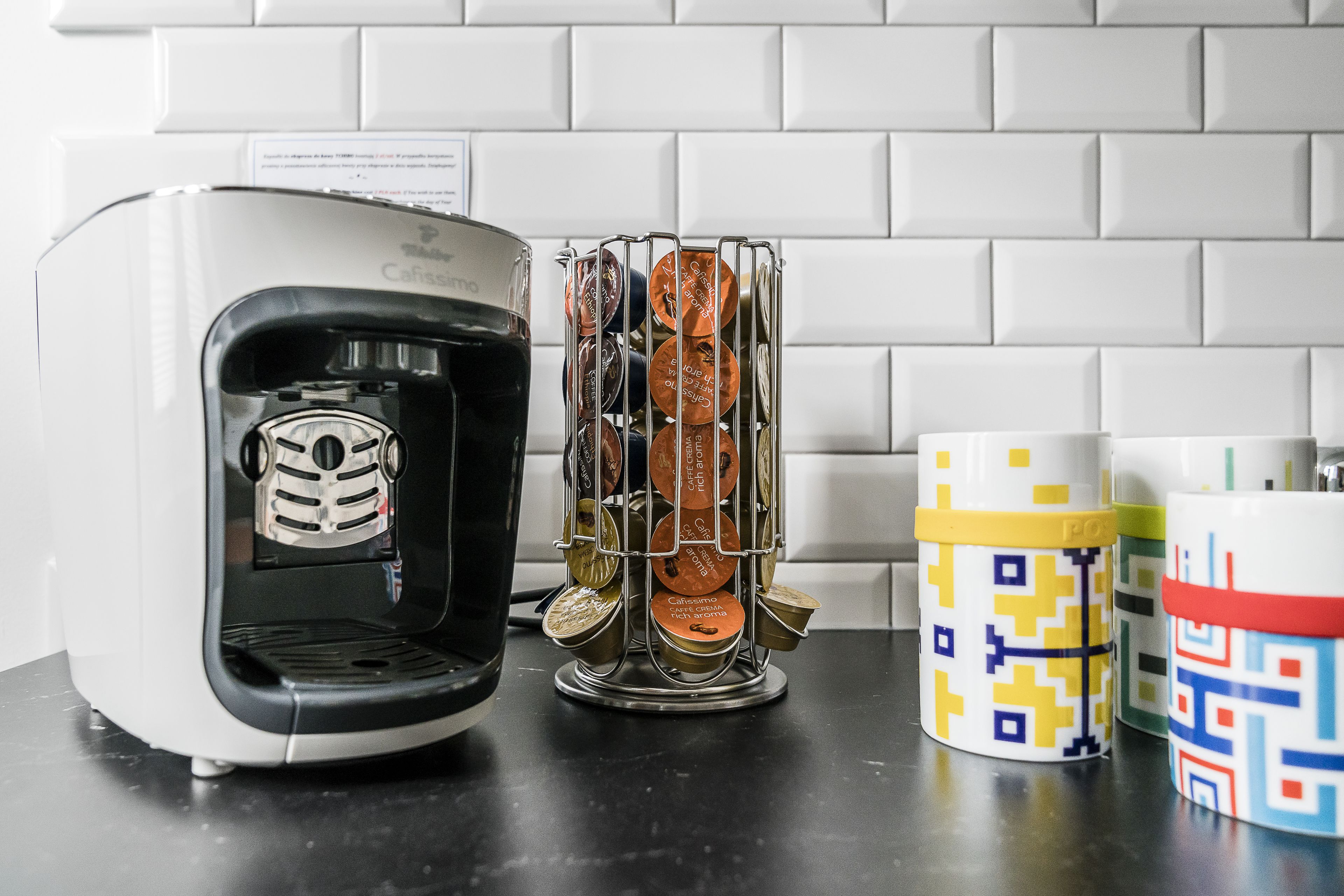 A coffee maker, a metal container with coffee capsules, and colorful mugs on a kitchen counter.