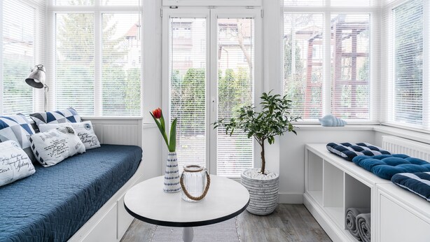 Una habitación luminosa con un sofá azul, una mesa redonda con un florero y una planta, y un asiento junto a la ventana con una planta y una lámpara.