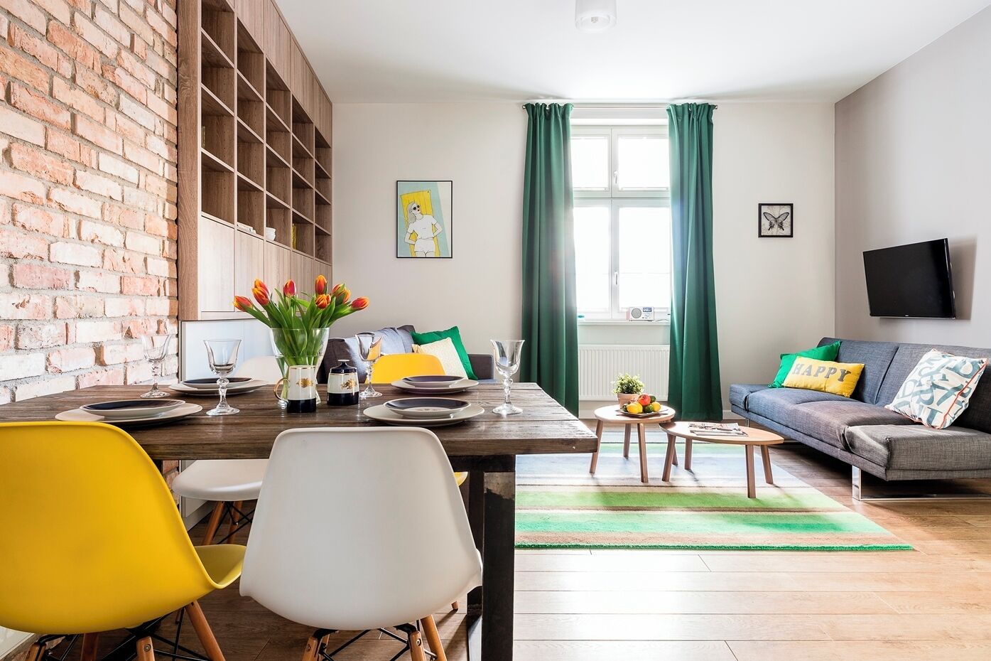 Un salon moderne avec un mur en briques, un coin repas avec une table en bois, des chaises jaunes et blanches, un canapé avec des coussins verts et un téléviseur à écran plat fixé au mur.