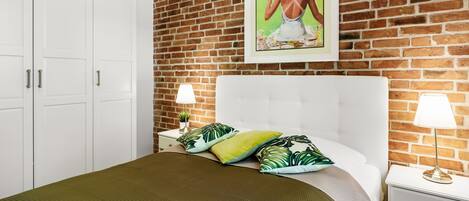 Un dormitorio con pared de ladrillo, cama con almohadas verdes y amarillas, cabecera blanca y un cuadro enmarcado sobre la cama.