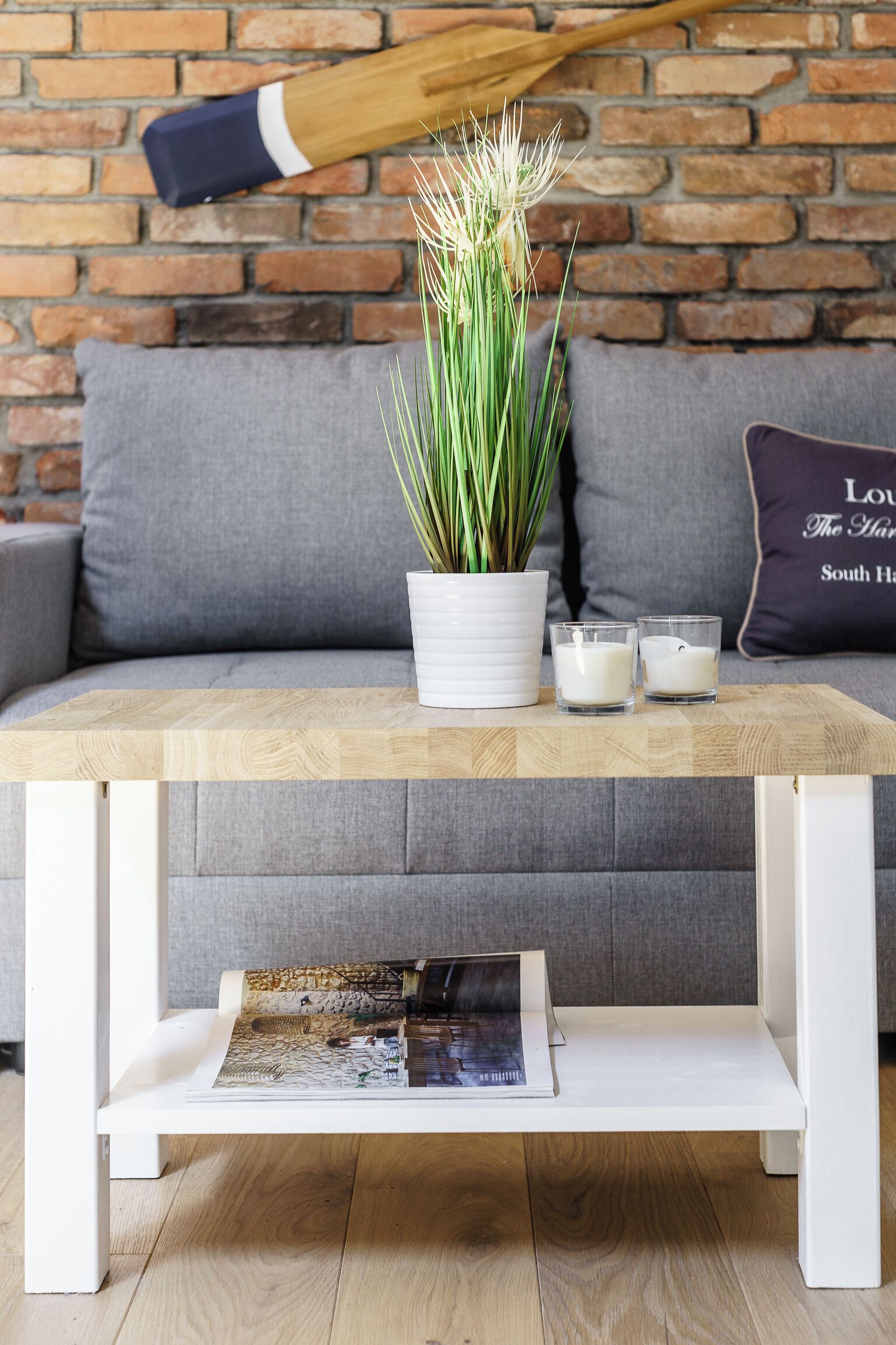 Une table basse en bois, ornée d’une plante en pot et de bougies, est placée sur un canapé gris, adossé à un mur de briques.