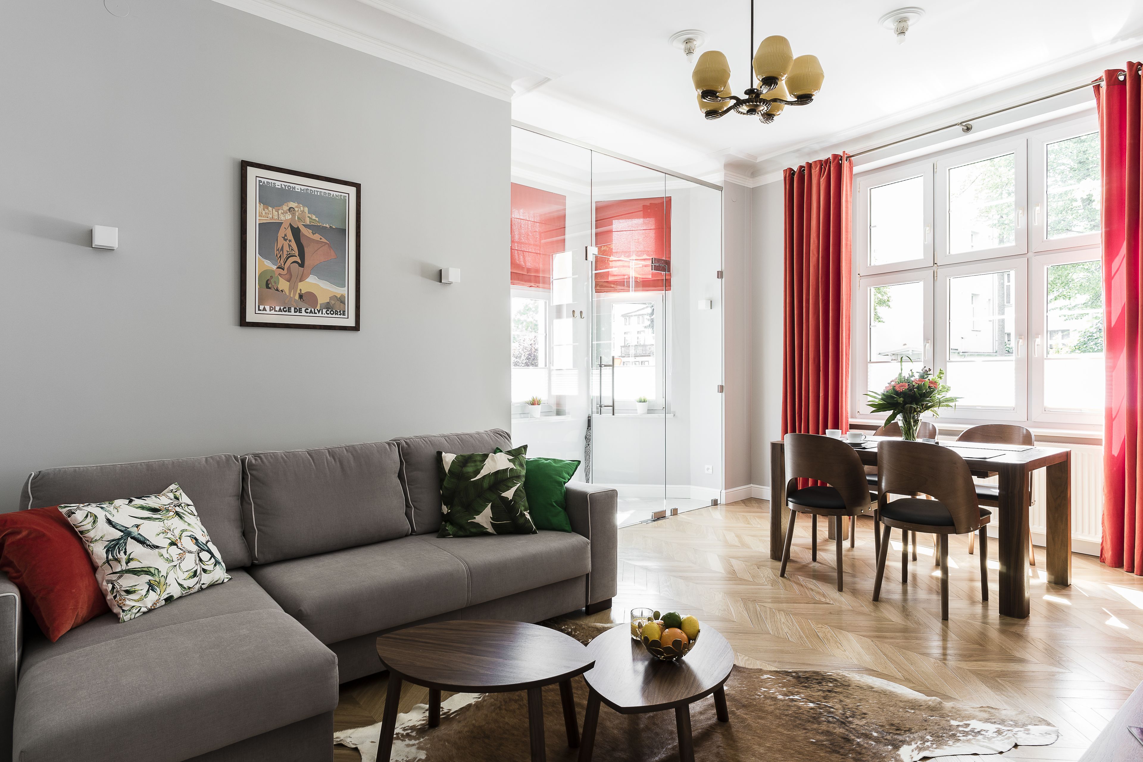 A modern living room with a grey sofa, a dining table with chairs, and a large window with red curtains.