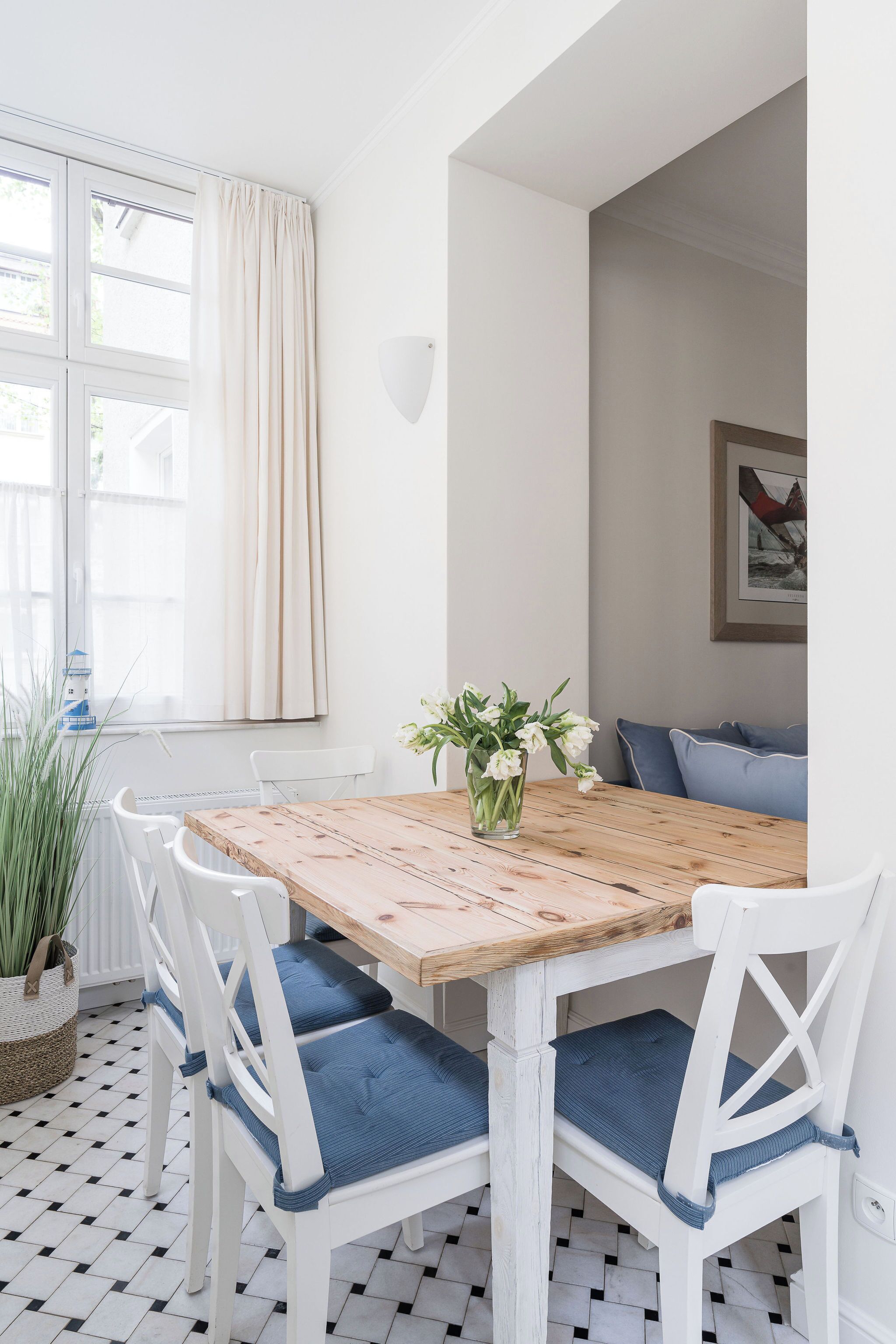 Un comedor con mesa de madera y sillas blancas con cojines azules, un florero con flores y una ventana con cortinas.