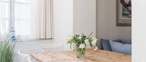 Un comedor con mesa de madera y sillas blancas con cojines azules, un florero con flores y una ventana con cortinas.