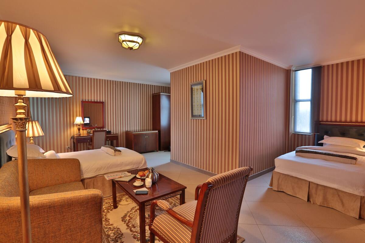 Quarto de hotel com duas camas, um espaço de estar com uma luminária, uma mesinha com bandeja e um espelho na parede.
