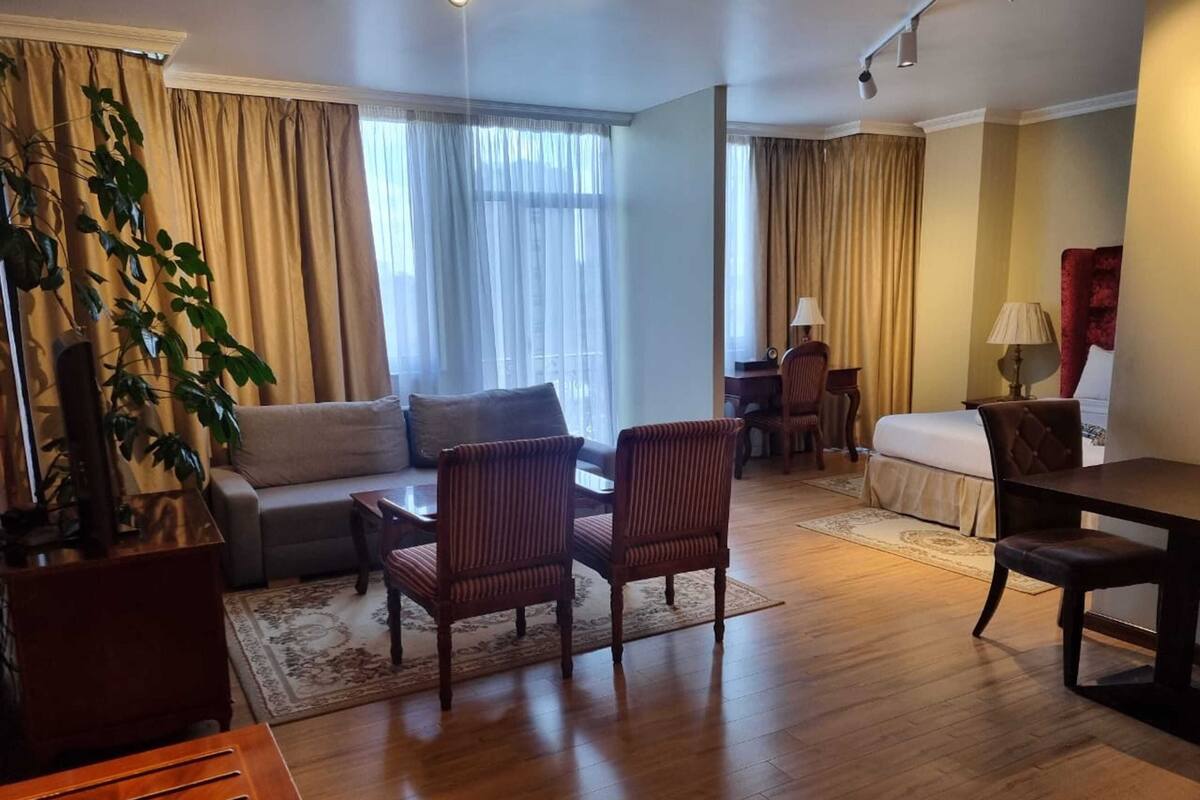 Quarto de hotel bem iluminado com sofá, duas poltronas, mesa de jantar, cama e escrivaninha.