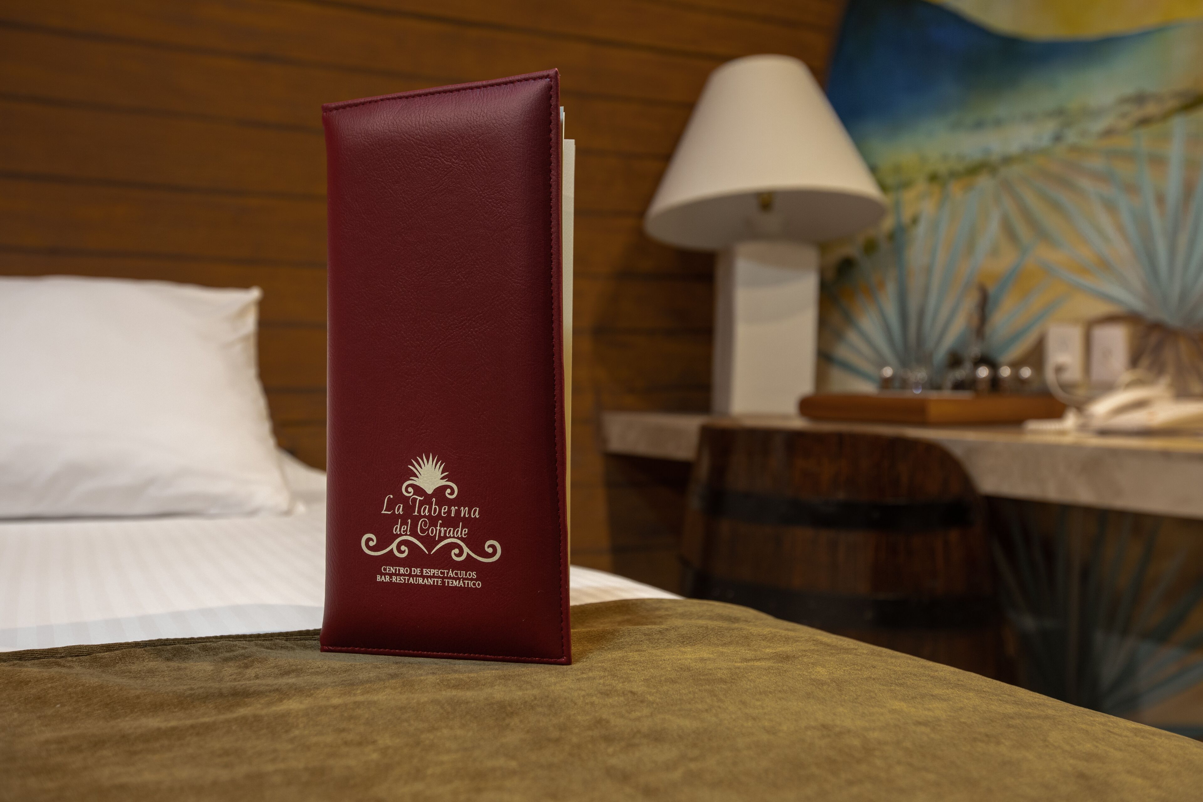 Un atril color burdeos con el texto "La Taberna del Cofrade" sobre la cama en una habitación de hotel.
