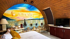 Un dormitorio con un mural de un atardecer, una cama con sábanas blancas y dos almohadas, una silla, un escritorio con una lámpara y un televisor.
