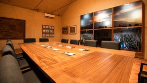 A conference room with a long wooden table, several chairs, framed pictures on the walls, and a large window with a view of mountains and a sunset.