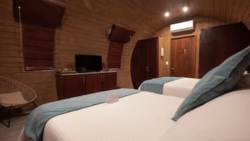 Quarto de hotel com painéis de madeira, duas camas com roupa de cama azul, uma televisão, um guarda-roupa e uma cadeira.
