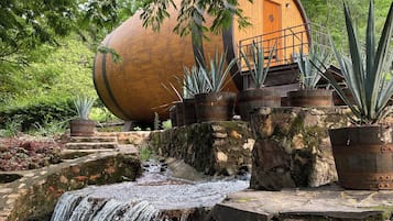 Una estructura rústica de barril de madera con puerta de madera, rodeada de plantas en macetas y una pequeña cascada.