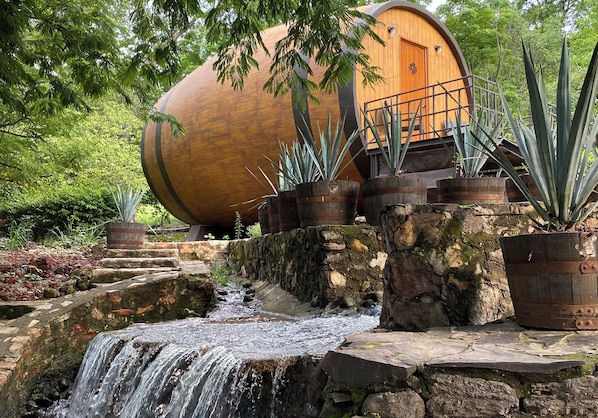 Une structure rustique en forme de tonneau, faite de bois, avec une porte en bois, entourée de plantes en pot et d’une petite cascade.