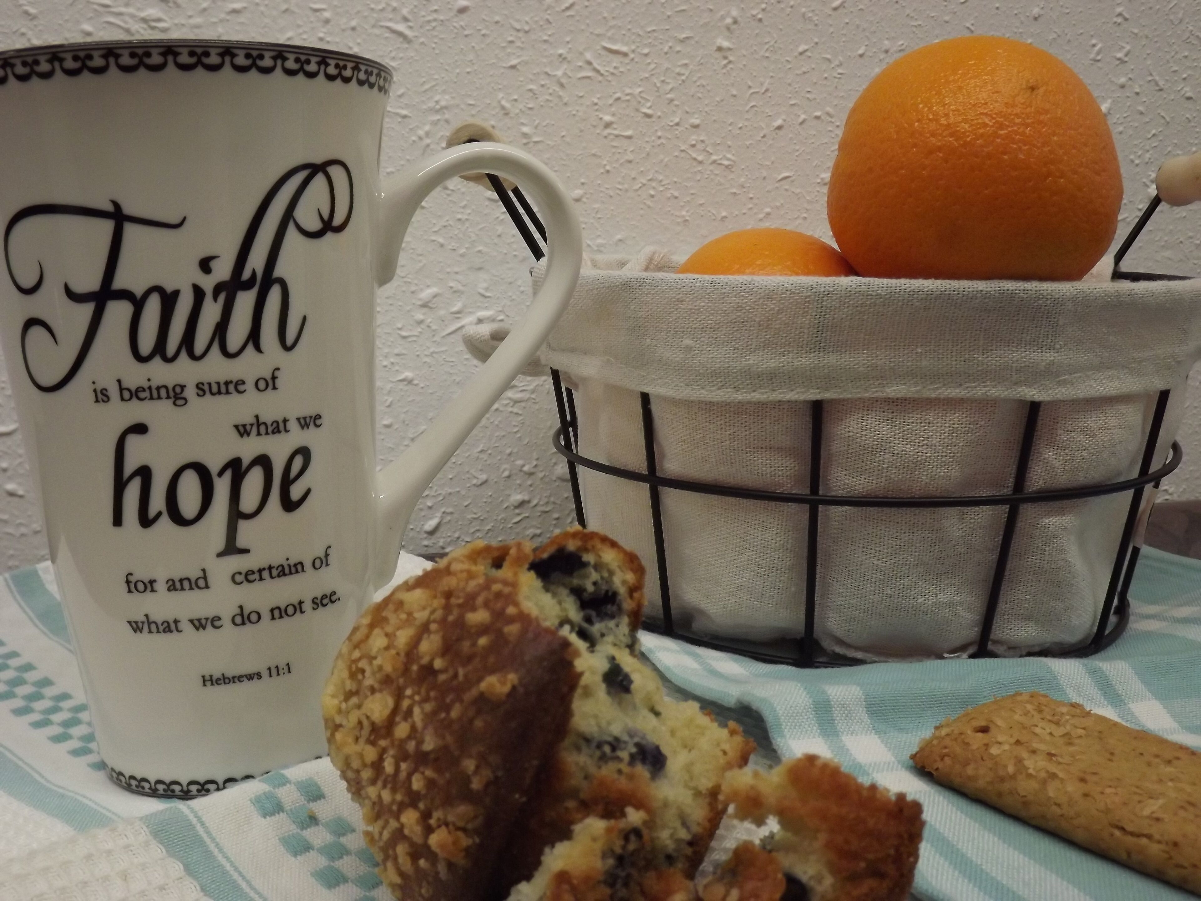 A white mug with black text, a basket of oranges, and a biscuit on a tablecloth.