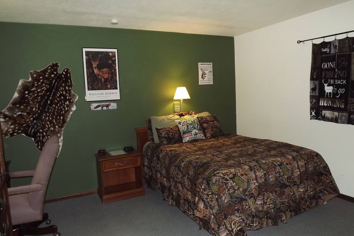 A bedroom with a green wall, a bed with a camouflage bedspread, a nightstand, a lamp, a poster of a deer, and a poster with hunting text.