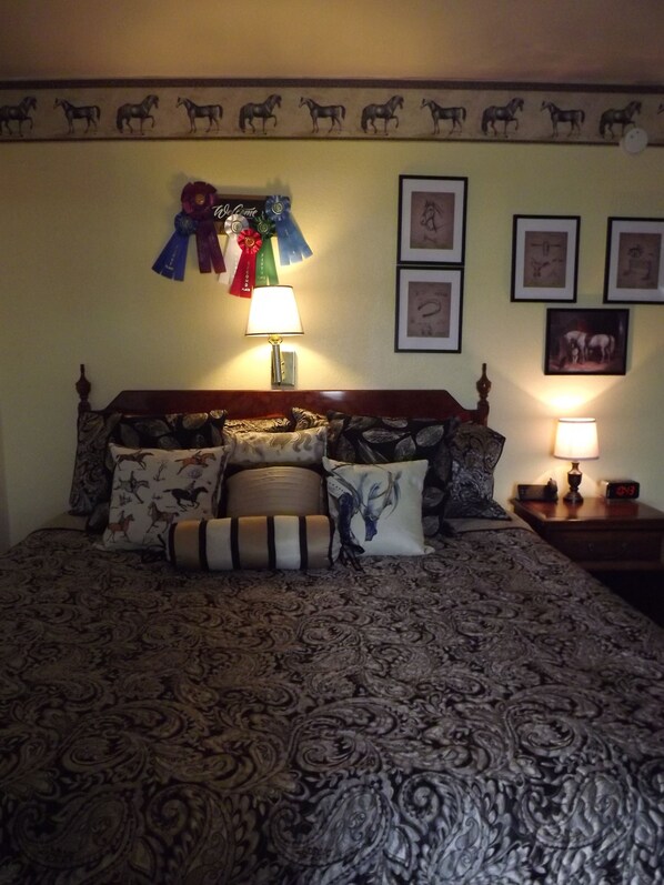Un dormitorio con colcha estampada, varias almohadas, una mesita de noche con lámpara, y decoración de pared que incluye cuadros enmarcados y un borde con motivos de caballos.