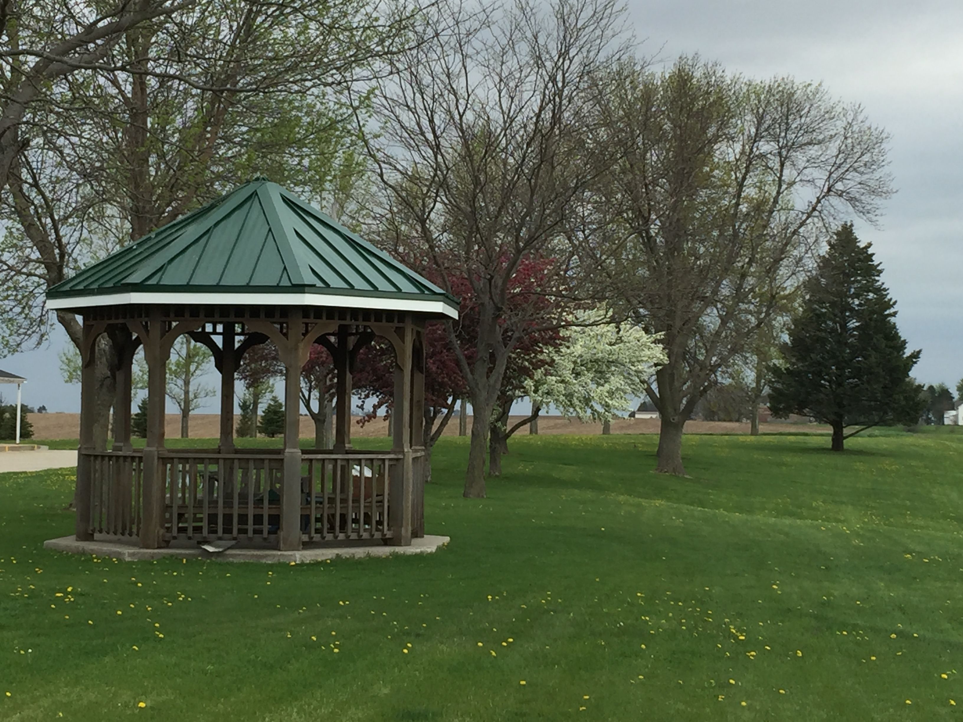 A gazebo with a green roof and wooden structure, surrounded by grass and trees.