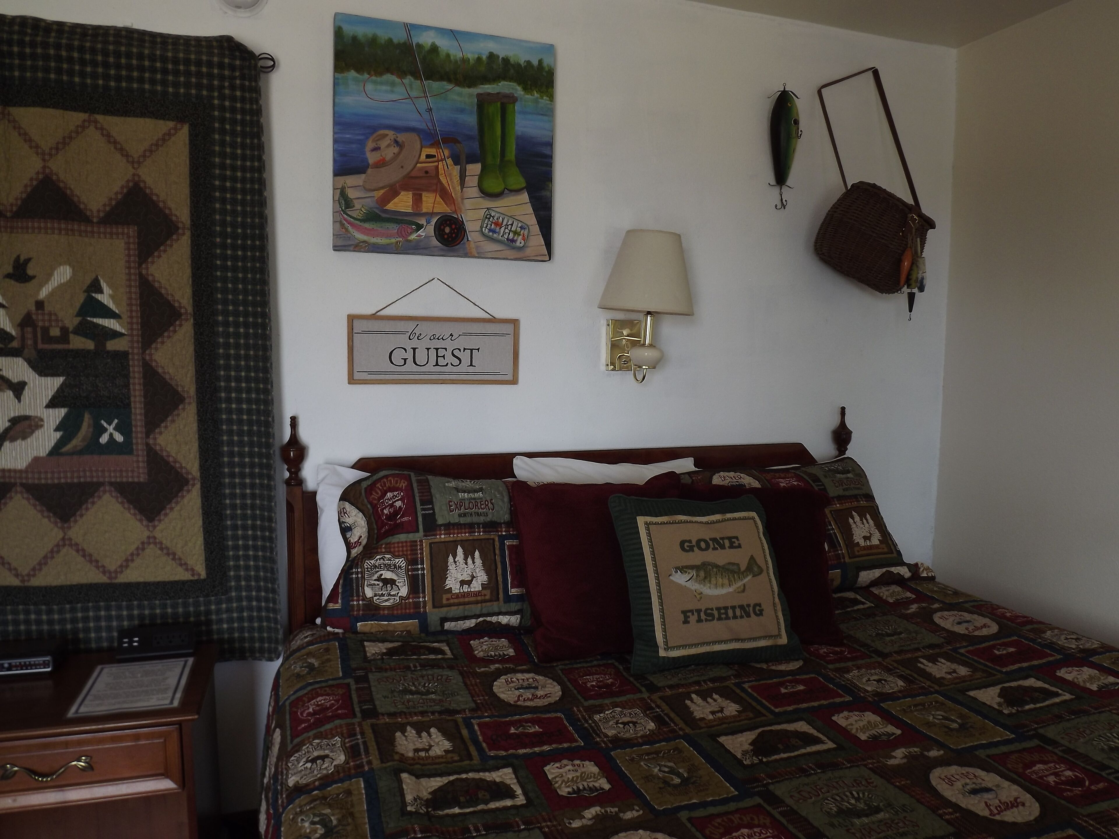 Quarto com colcha acolchoada exibindo temas da natureza selvagem e atividades ao ar livre, um letreiro com a frase 'be our guest', um abajur e uma almofada com tema de pesca.