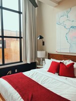 Un cuarto de hotel con un ventanal grande, una cama con ropa de cama blanca y roja, una mesita de noche con una lámpara y un mapa en la pared.
