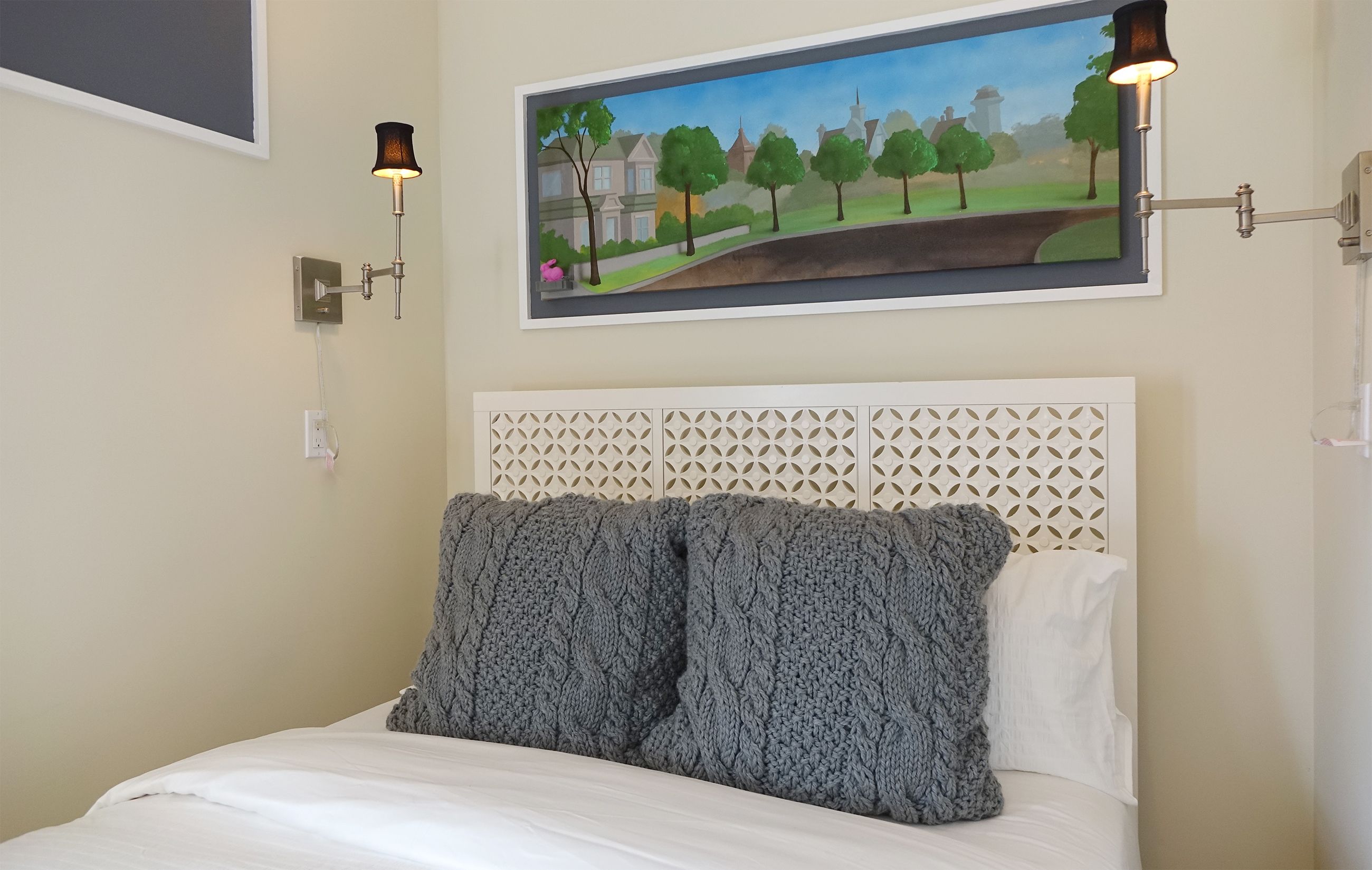 Un dormitorio con cabecera blanca, un cuadro de un paisaje y dos lámparas de pared.