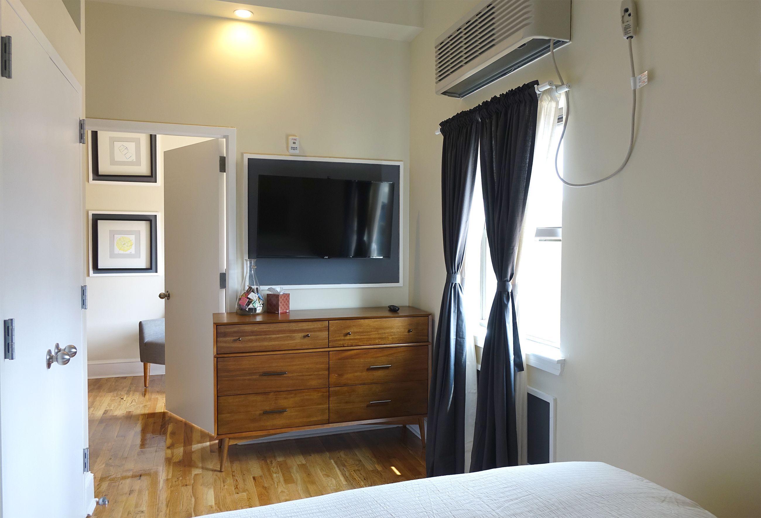 A bedroom with a bed, a TV mounted on a wooden dresser, a window with curtains, and a door leading to another room.