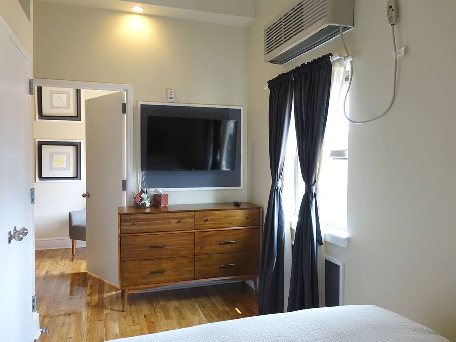 A bedroom with a bed, a TV mounted on a wooden dresser, a window with curtains, and a door leading to another room.