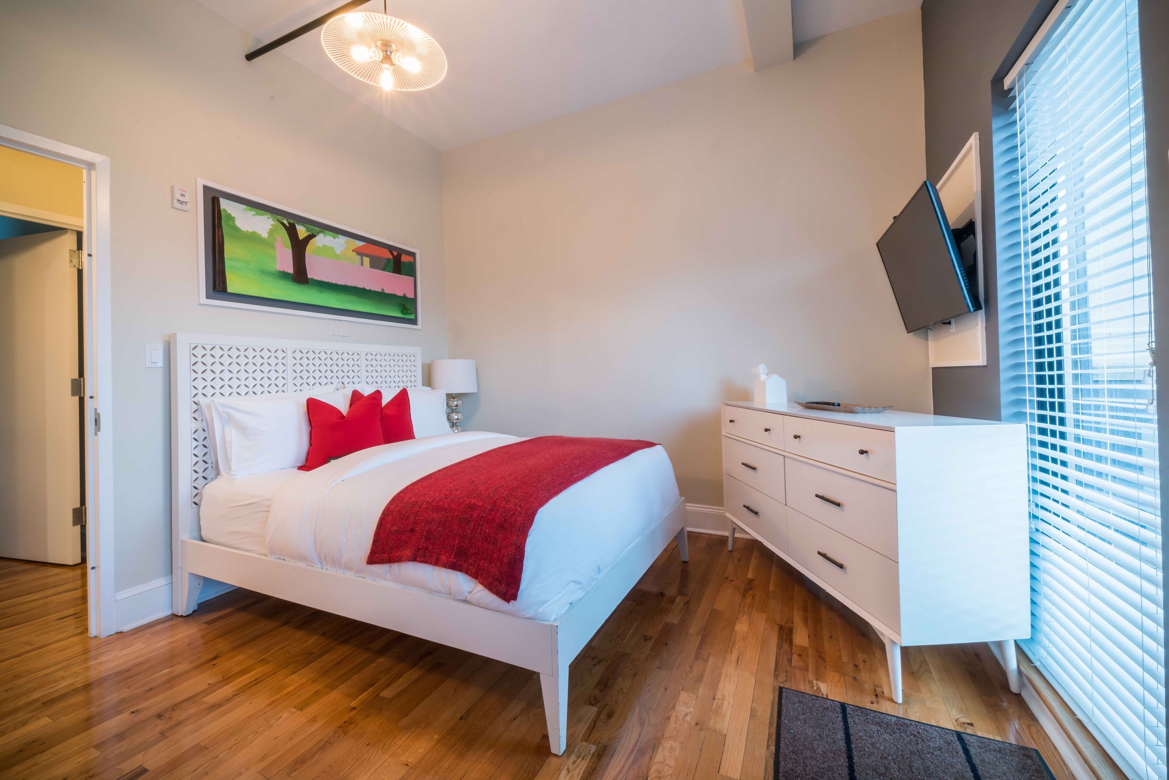 A bedroom with a white bed, red pillows, a white dresser, a TV on the wall, and a painting above the bed.