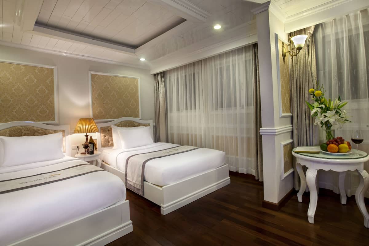 Quarto de hotel com duas camas, mesa de cabeceira com abajur, pequena mesa com frutas e uma taça de vinho, e uma grande janela com cortinas leves.