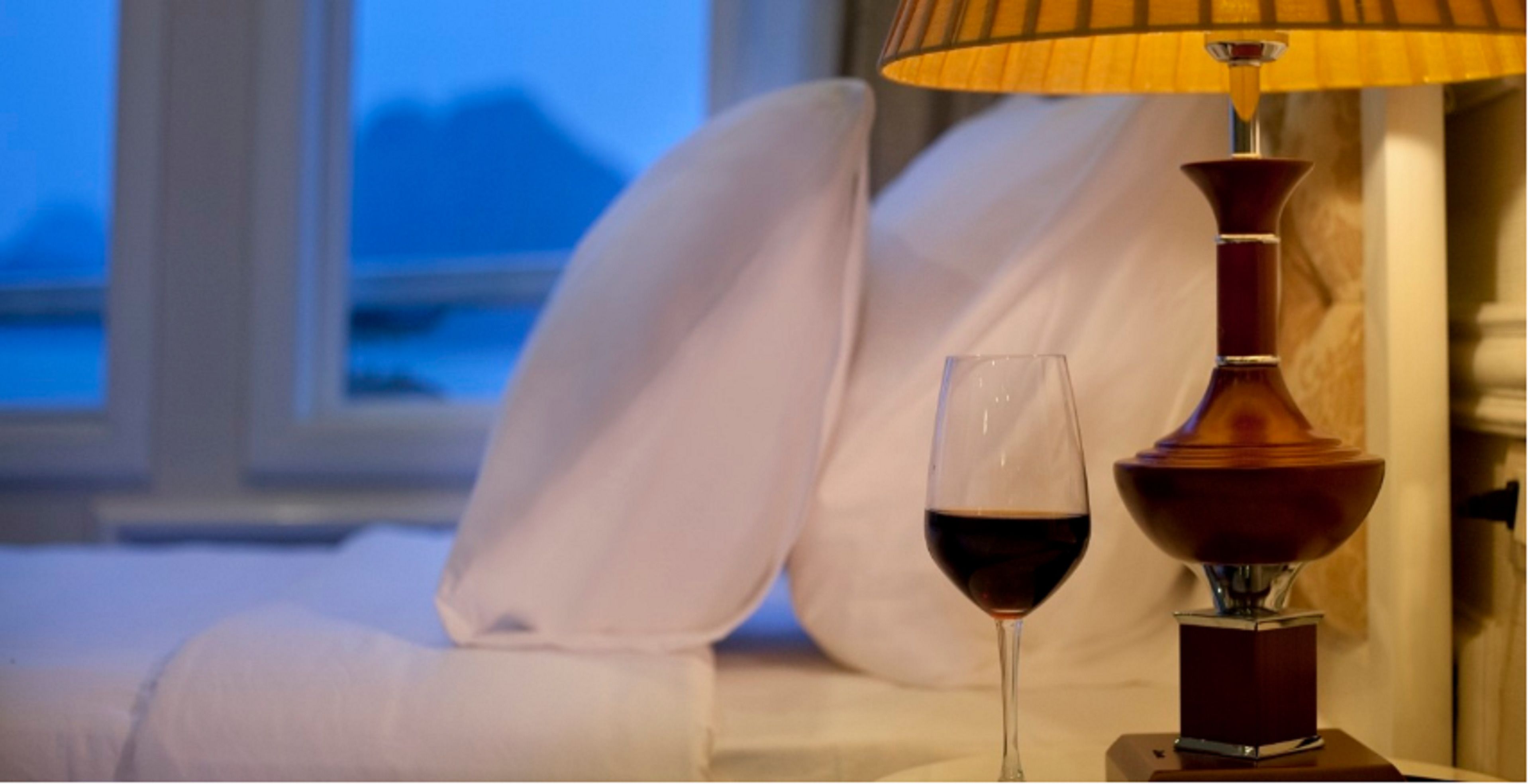 Un letto con lenzuola bianche, un bicchiere di vino rosso e una lampada da tavolo con base in legno e paralume giallo.