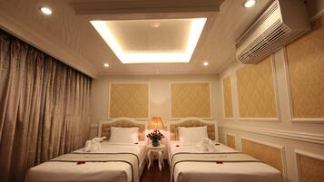 A hotel room with two beds, a ceiling light, an air conditioner, and a small table with a lamp and flowers.