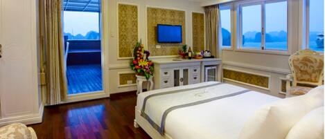 A spacious bedroom with a large bed, a television on the wall, a desk with a chair, and a view of the sea through the window.