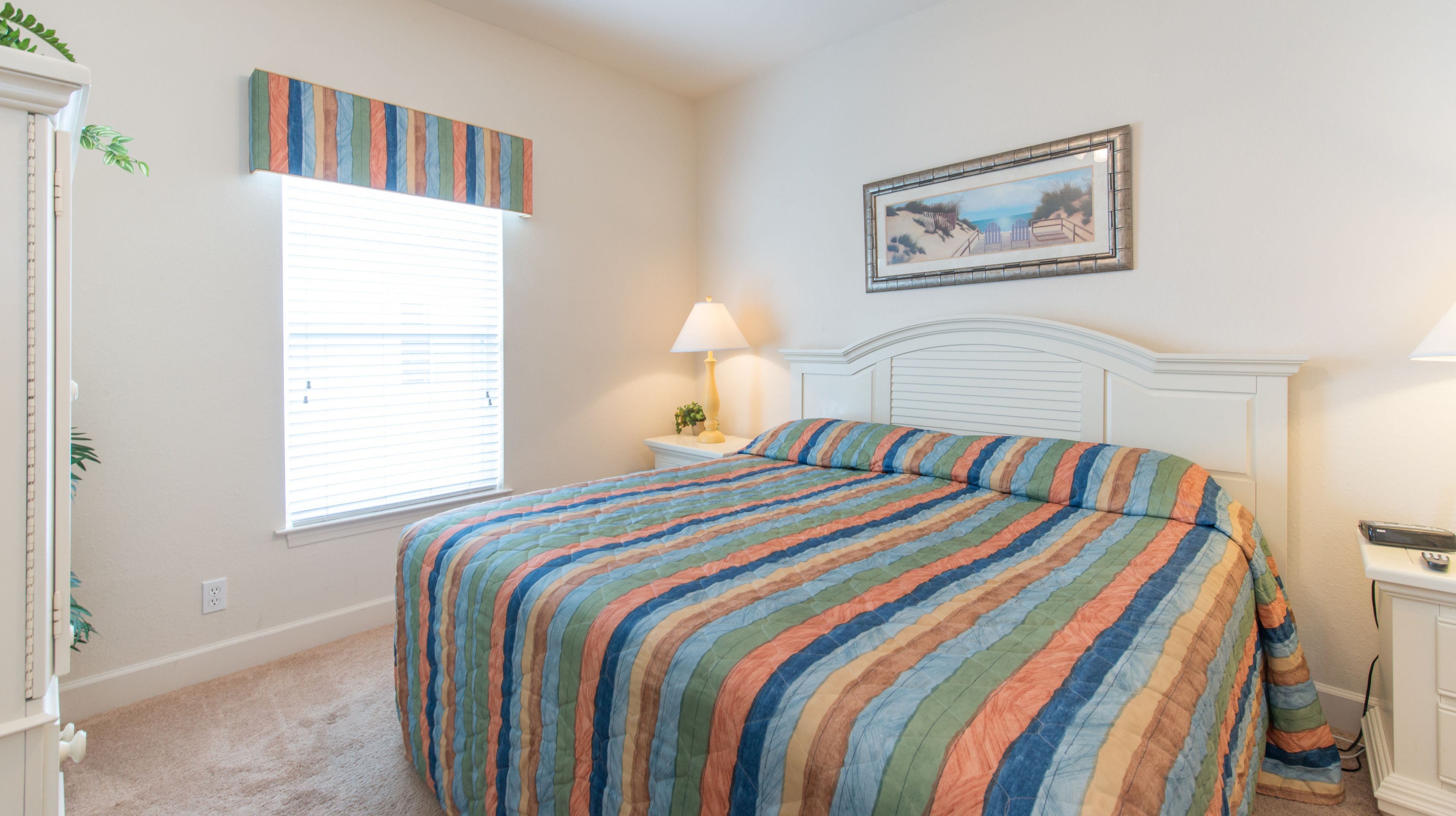 A bedroom with a striped bedspread, a window with blinds, a bedside table with a lamp, and a framed picture on the wall.