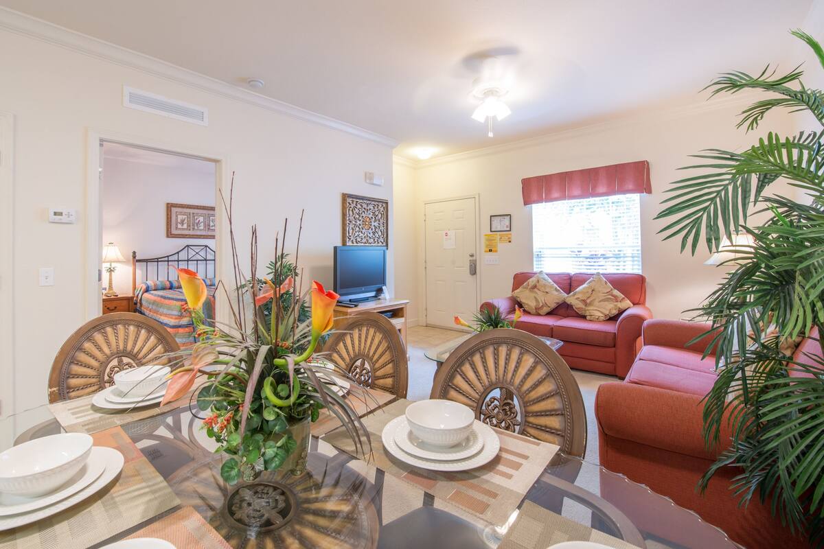 A dining area with a round table set for four, a television in the background, and a red sofa in the living room.