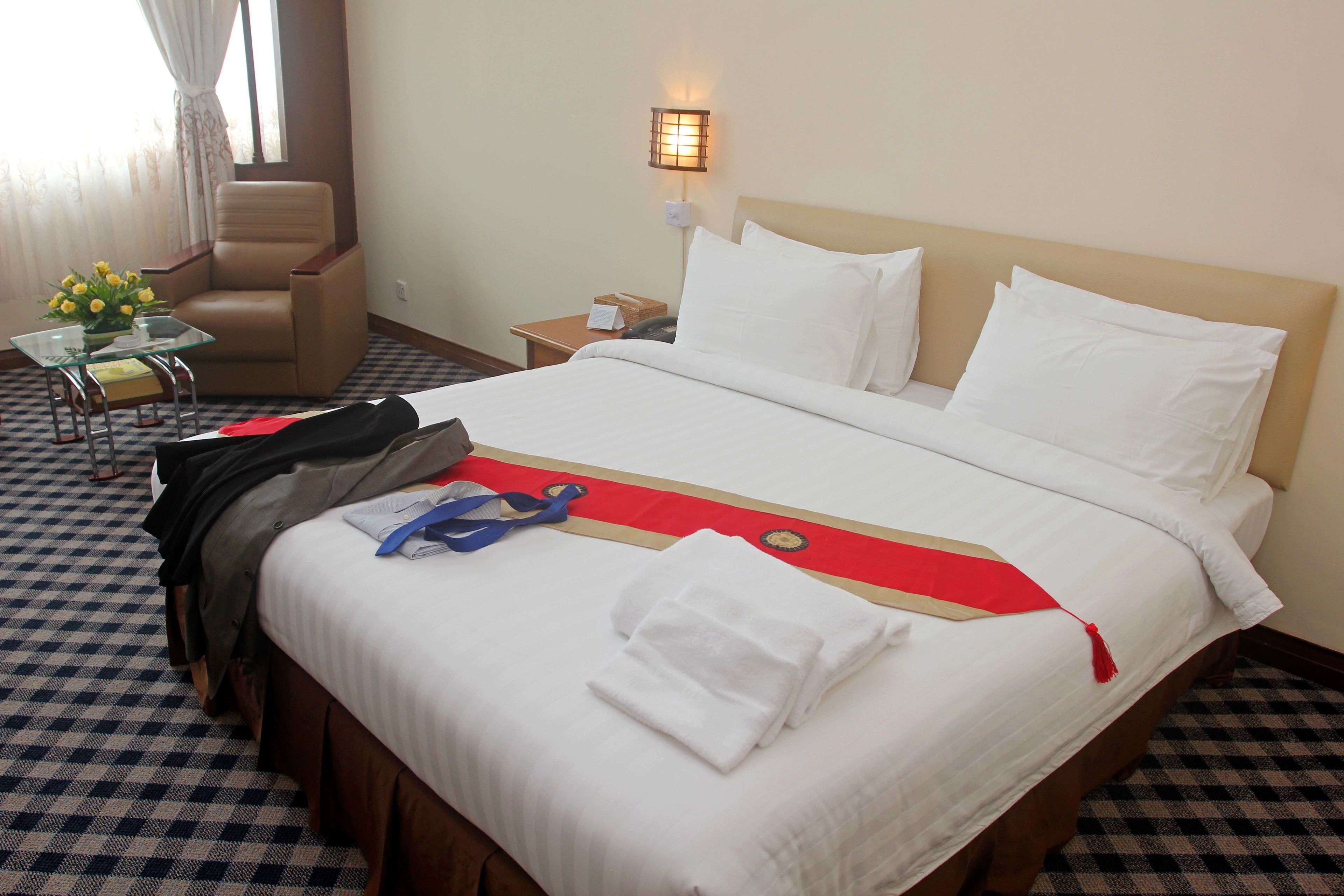 A hotel room with a checkered floor, a bed with white linens, a chair, a small table with a vase of flowers, and a wall-mounted lamp.