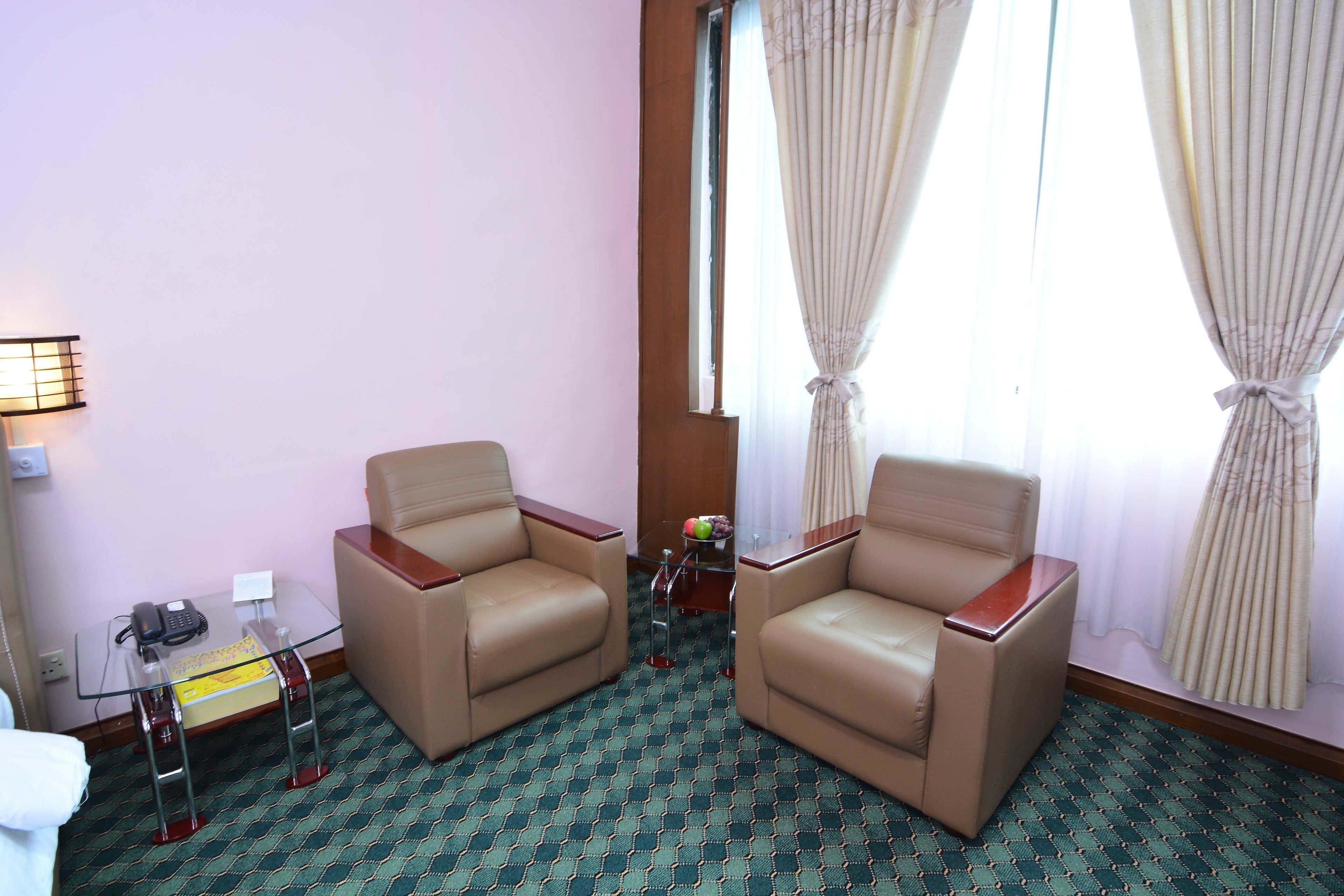 Habitación de hotel con dos sillones de cuero, una mesita de vidrio con teléfono y una caja pequeña, una lámpara y una ventana con cortinas.
