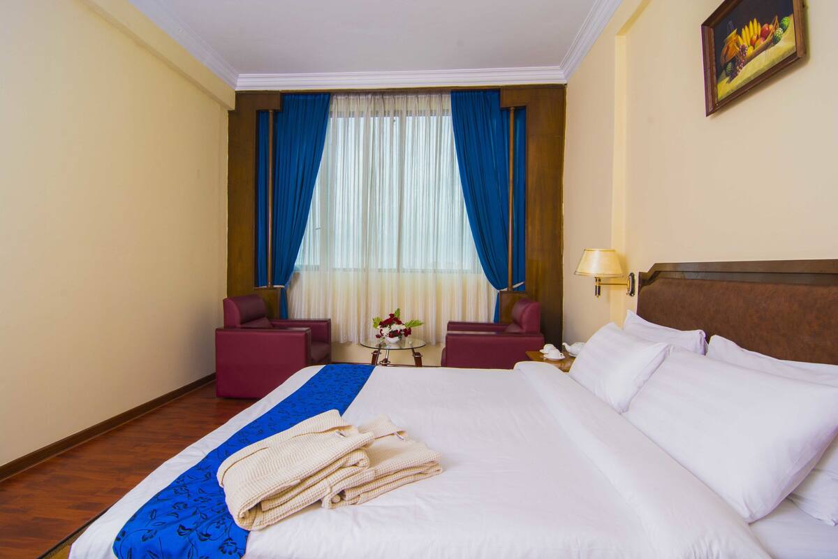 Une chambre d’hôtel avec un grand lit, deux fauteuils, une petite table avec un vase et un tableau au mur.