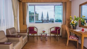 Habitación de hotel con un ventanal grande, dos sillas rojas, una mesa de vidrio con frutero, un escritorio de madera y un espejo.